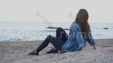 美丽的年轻女子坐在海滩上放松，在温暖的夏天晚上欣赏海天一色的景色。